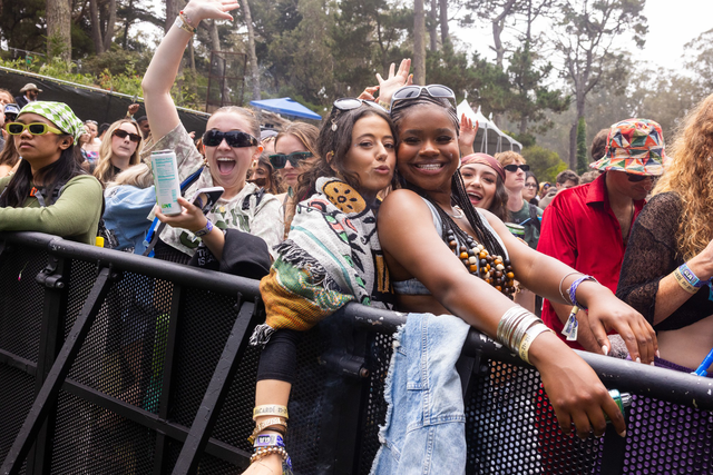 A diverse group of people stand behind a metal barrier at an outdoor event, smiling, posing, and enjoying themselves in casual, colorful clothing.